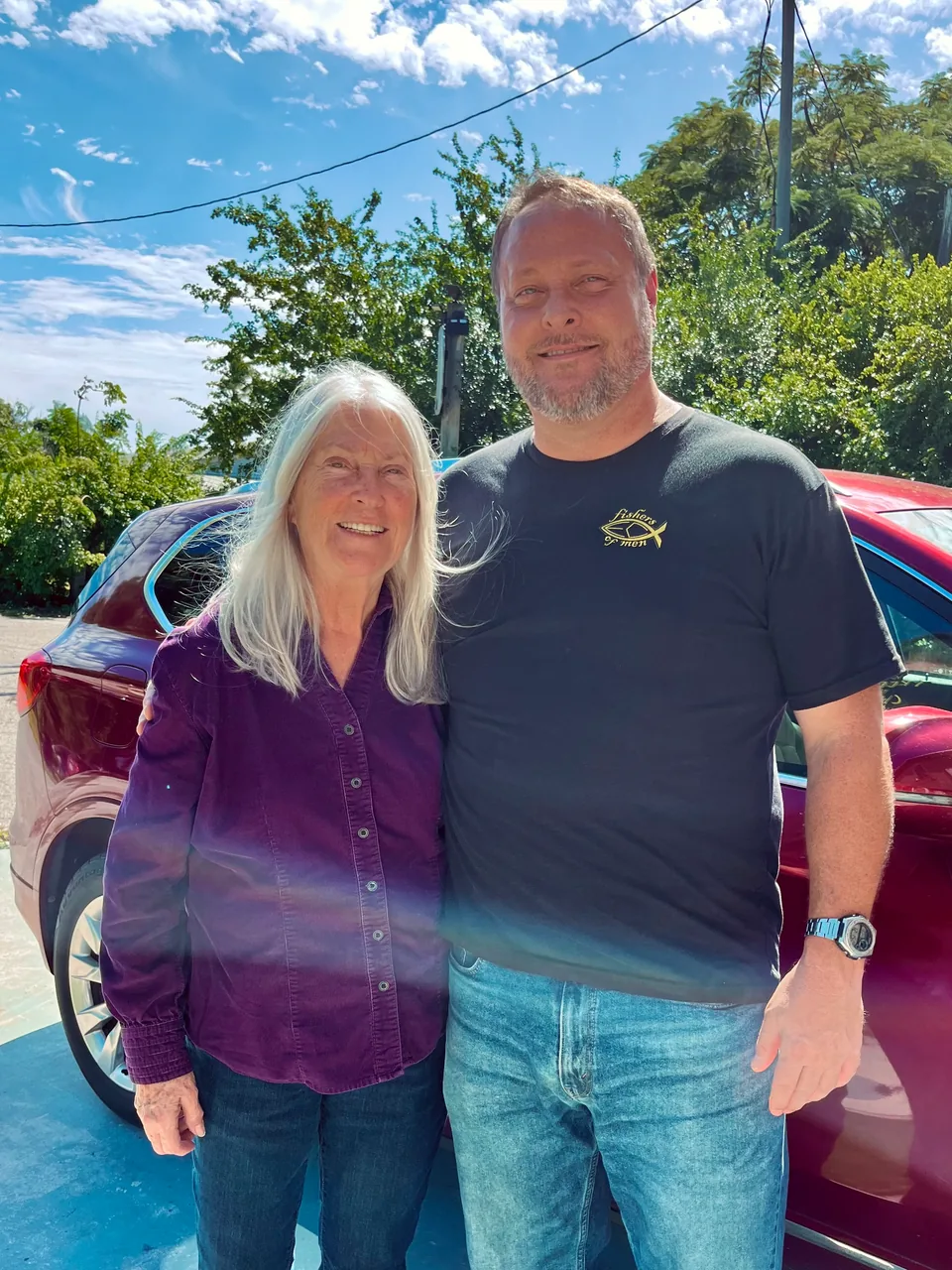 A woman and man stand together beside a red car outdoors on a sunny day.